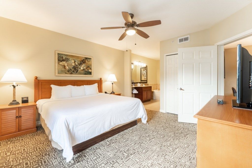 Une chambre d’hôtel comprenant un lit, deux lampes de chevet, un ventilateur de plafond, une table de chevet en bois, une commode, un téléviseur et une moquette à motifs.