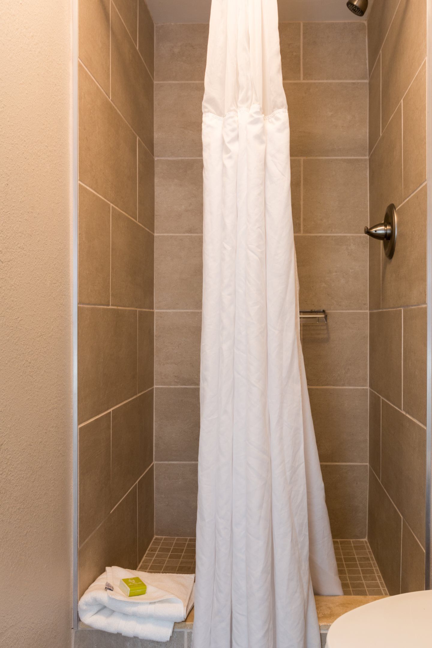 A shower area with beige tiles, a white shower curtain, and a towel with a green bar of soap on the floor.