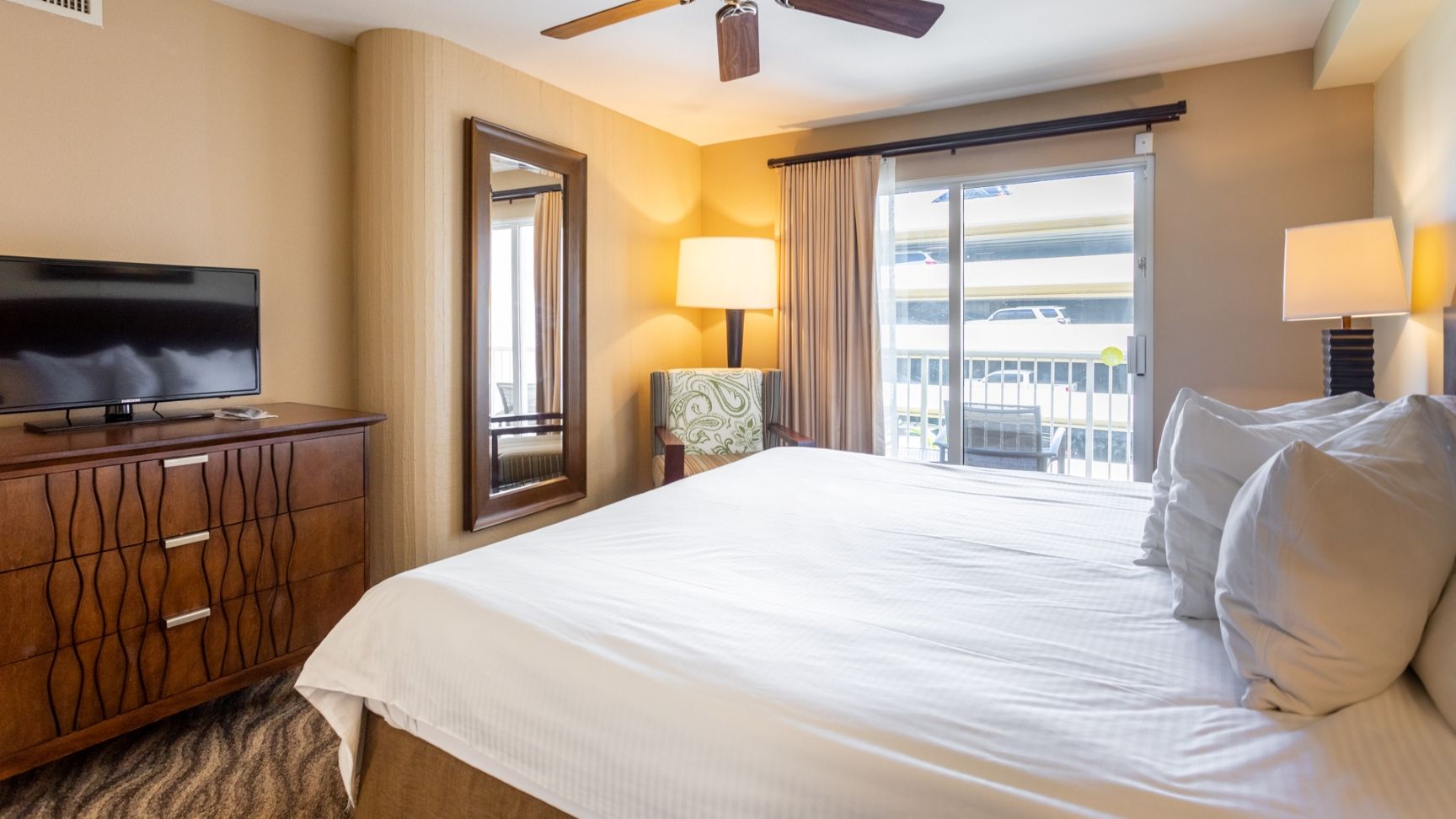 A hotel room with a bed, a television on a wooden dresser, a chair, and a balcony with a view.