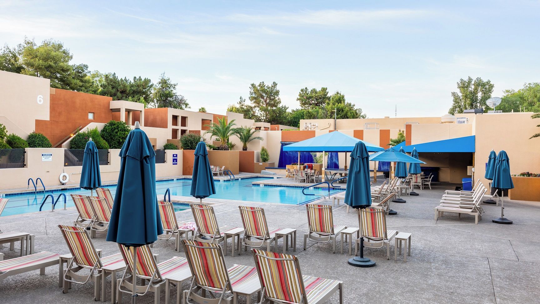 A pool area with lounge chairs and umbrellas, a clear sky, and a building with a number '6' sign.