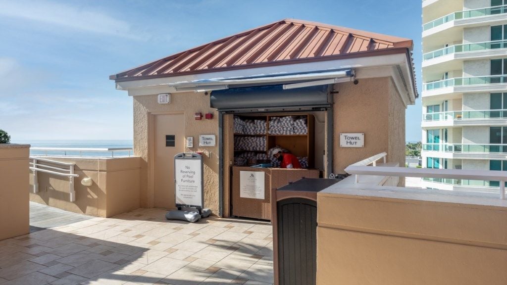 Ein Hotel direkt am Strand mit Bademäntelstand, Schild zur Reservierung von Poolliegen und Blick auf das Meer.