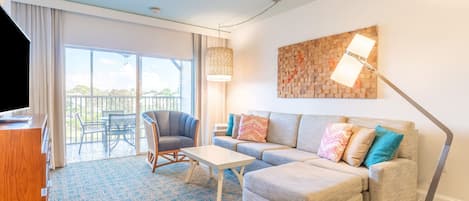 A modern living room with a sofa, armchair, and coffee table. There is a balcony with outdoor furniture and a large window.