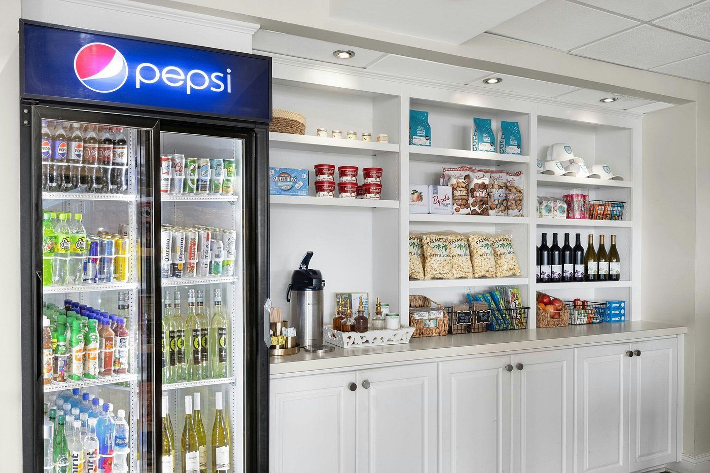 A beverage refrigerator stocked with various drinks, a coffee machine, and shelves with snacks and bottled water.
