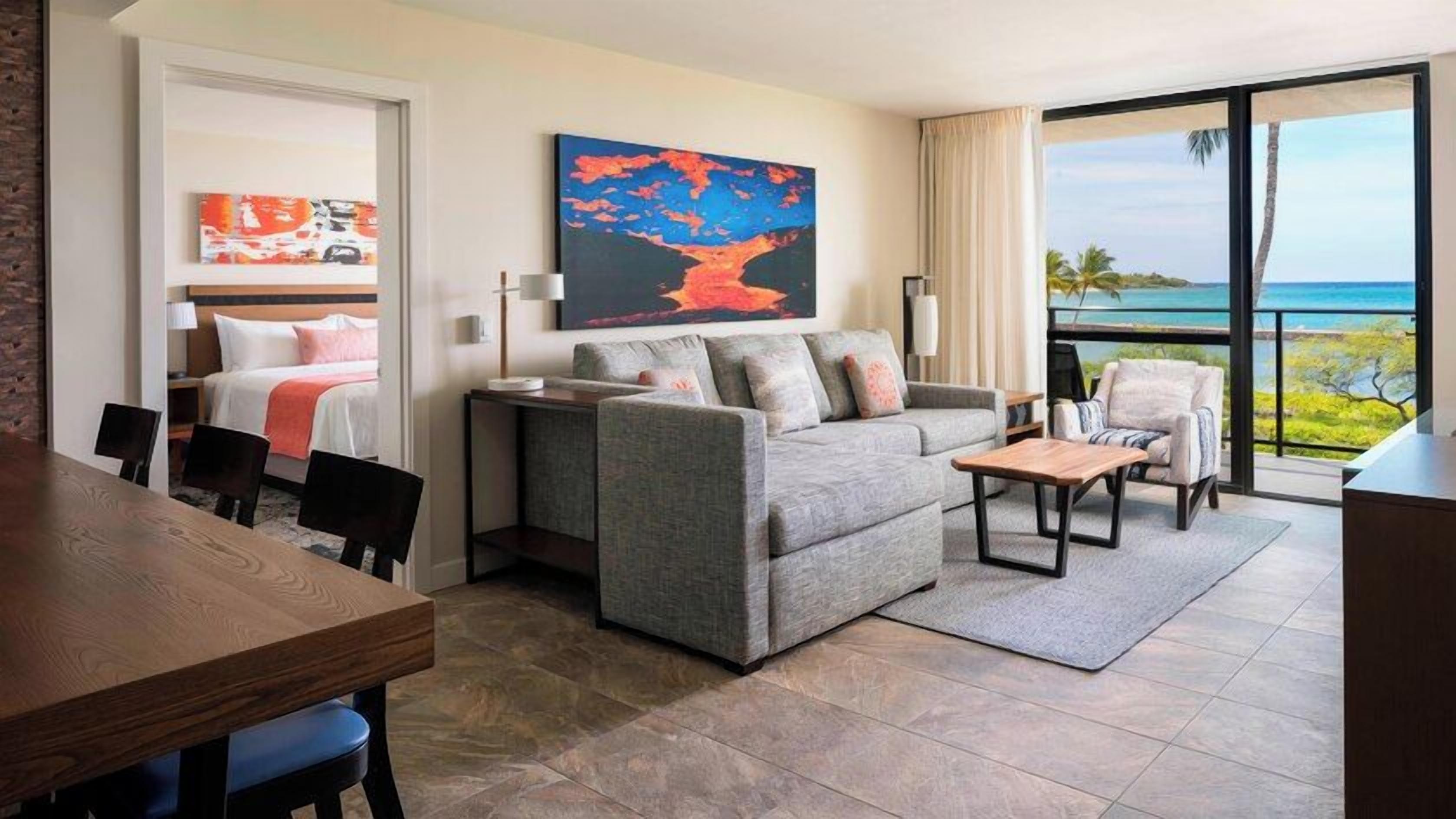 A modern hotel room with a balcony, a sofa, a dining table, and a bed with a view of the ocean.