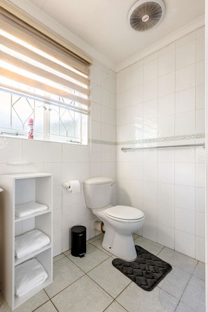 Un baño con inodoro, un estante con toallas, un cesto de basura negro y una ventana con persianas.