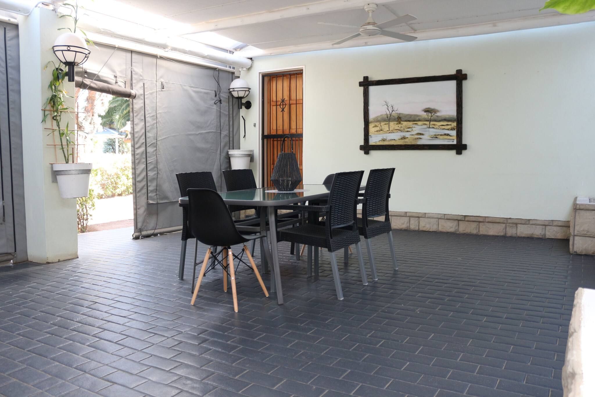 Een terras buiten met een tafel, stoelen, een plafondventilator en een gekaderd schilderij aan de muur.