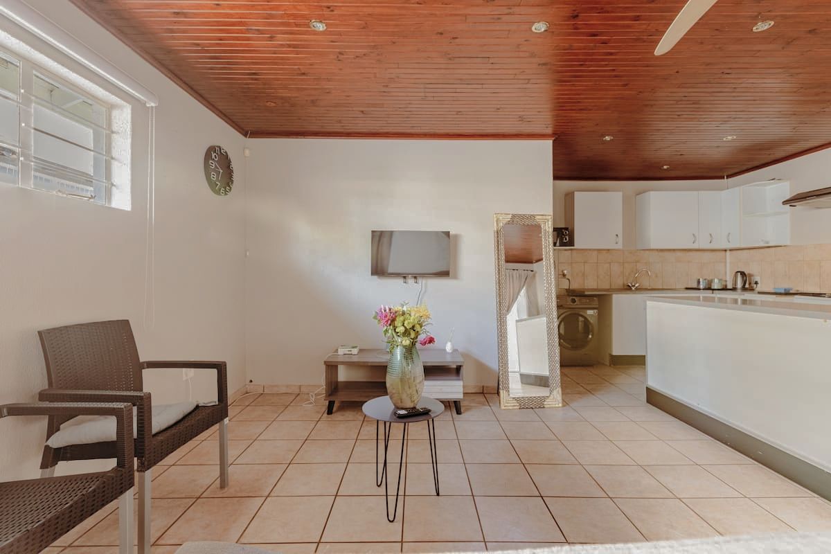 Ein Zimmer mit Holzboden, einem Fernseher, einem kleinen Tisch mit Blumenvase, einem Stuhl und einer Küchenzeile inklusive Schränken und Waschmaschine.