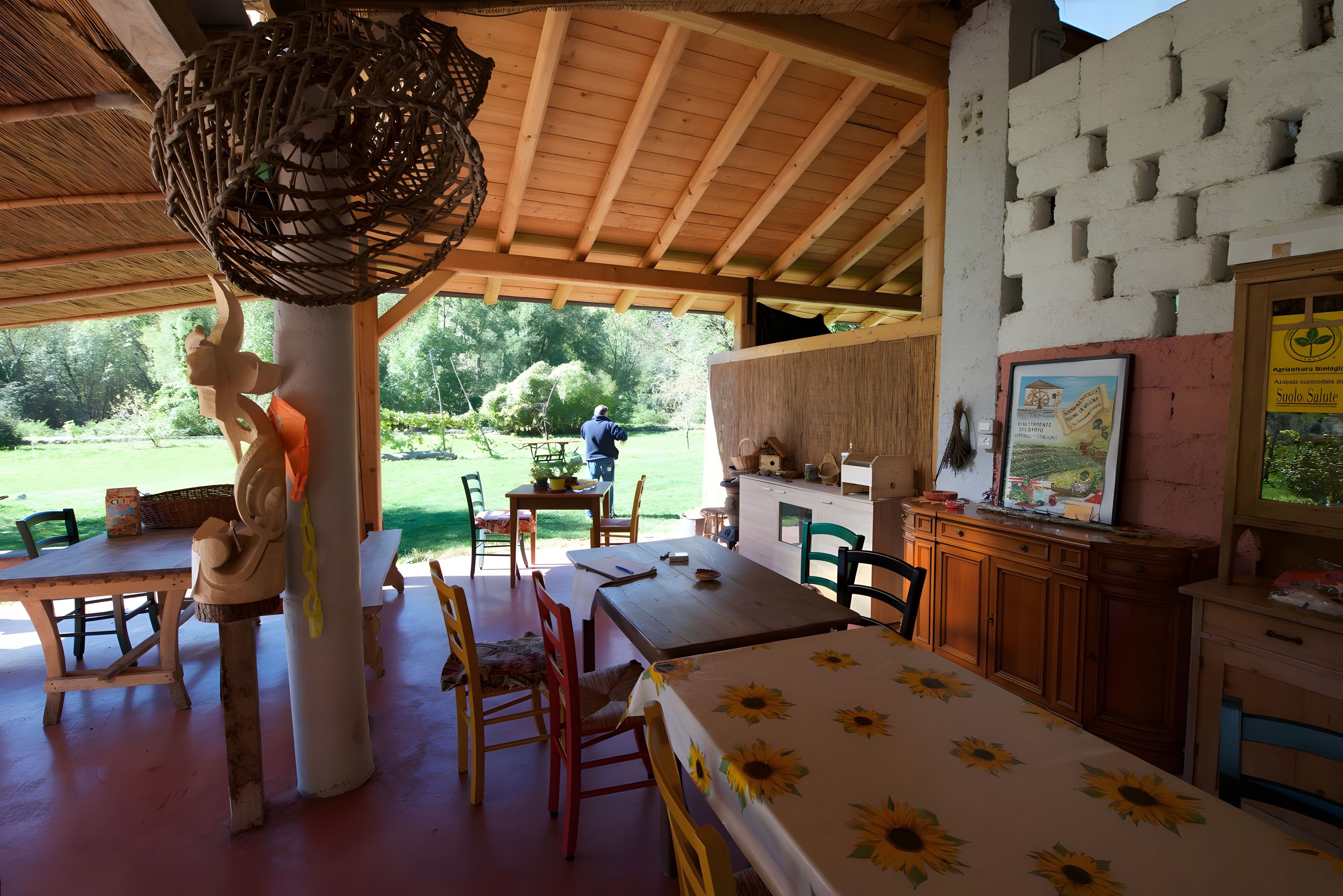 An outdoor dining area with wooden tables and chairs, a hanging light fixture, and a view of the garden.