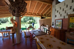 Un área de comedor al aire libre con mesas y sillas de madera, una lámpara colgante y vistas al jardín.
