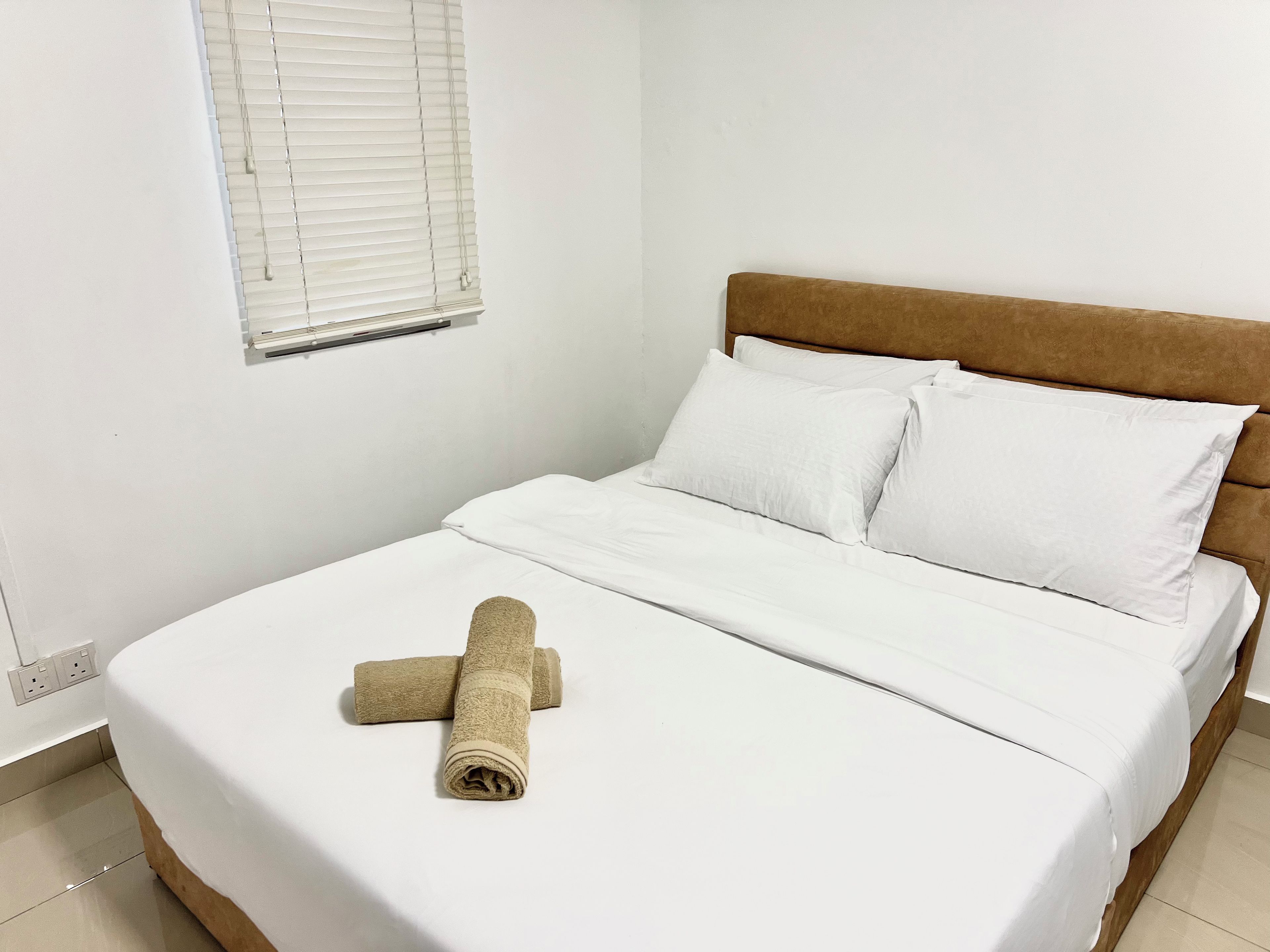 A single bed with white bedding and a brown headboard, a rolled-up beige towel, and a window with closed blinds.