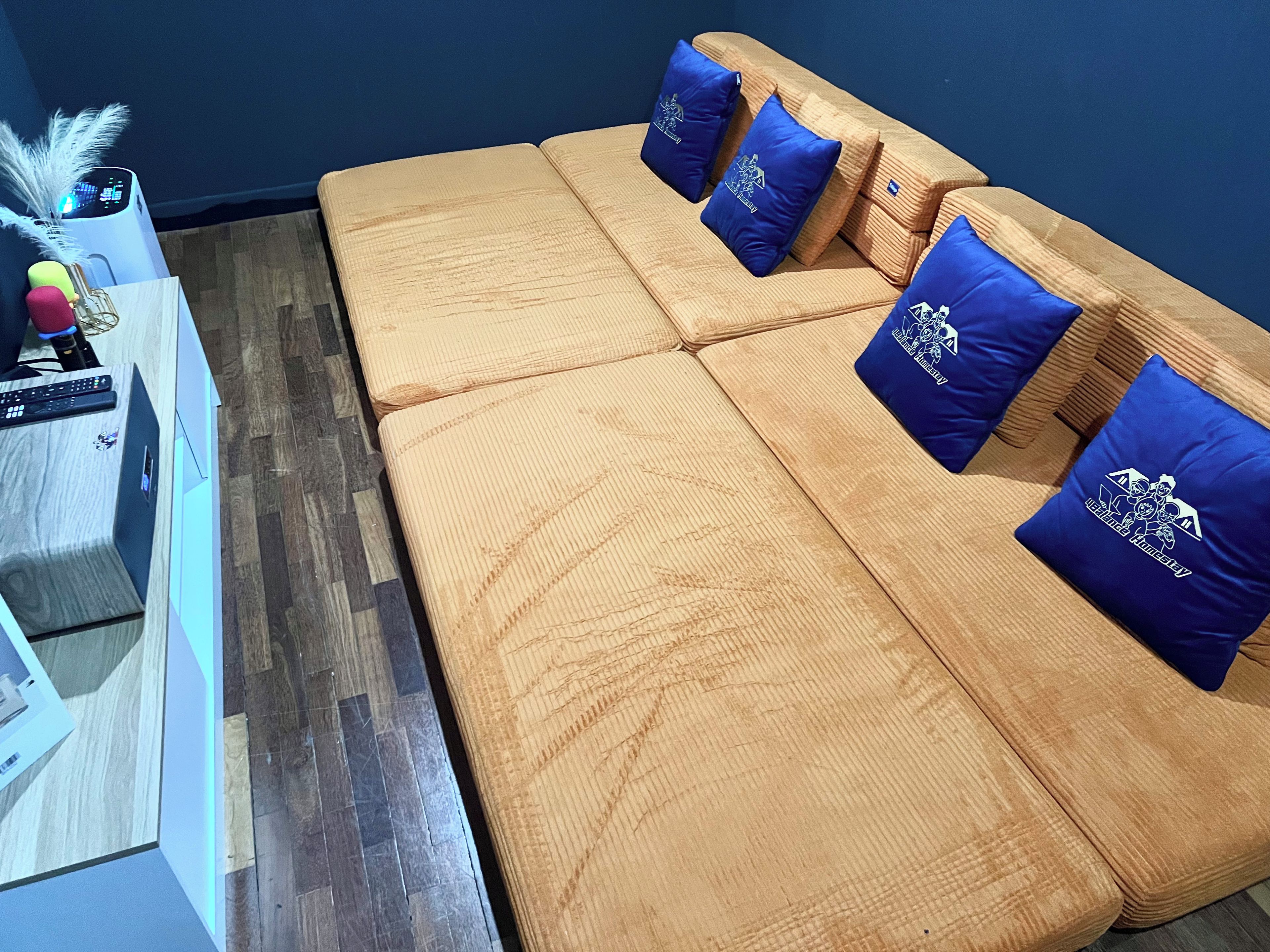 A room with a wooden sofa and blue cushions, a small table with a vase and a remote control, and a wall-mounted speaker.