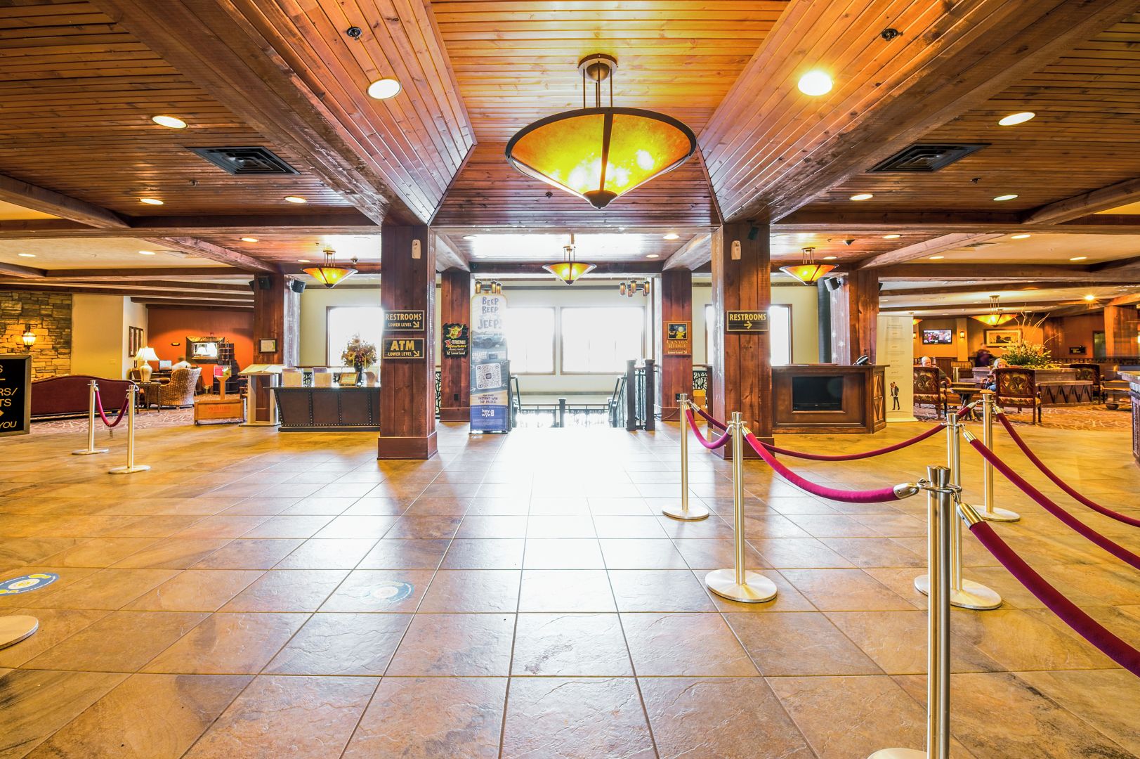 A spacious lobby with a high ceiling, wooden beams, and a central chandelier. There are signs for restrooms and an ATM.