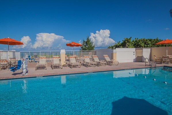 Ein Swimmingpool mit Liegestühlen und Sonnenschirmen, umgeben von einem weißen Zaun und blauem Himmel.