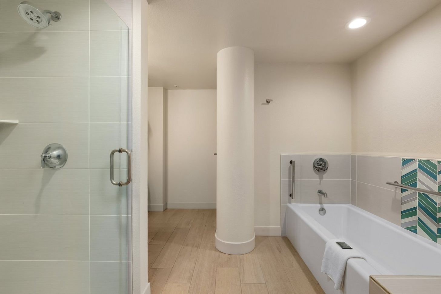 Un baño moderno con cabina de ducha de vidrio, bañera con salpicadero de azulejos blancos y verdes, y suelo de madera.