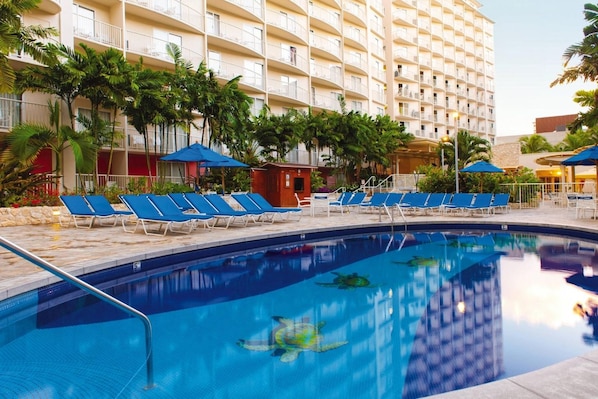 Una piscina de hotel con sillas reclinables azules, palmeras y un edificio de varios pisos al fondo.