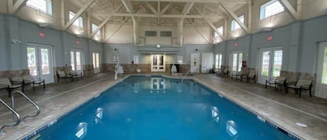 An indoor swimming pool with a high ceiling and tiled floor, surrounded by chairs and a door in the center.