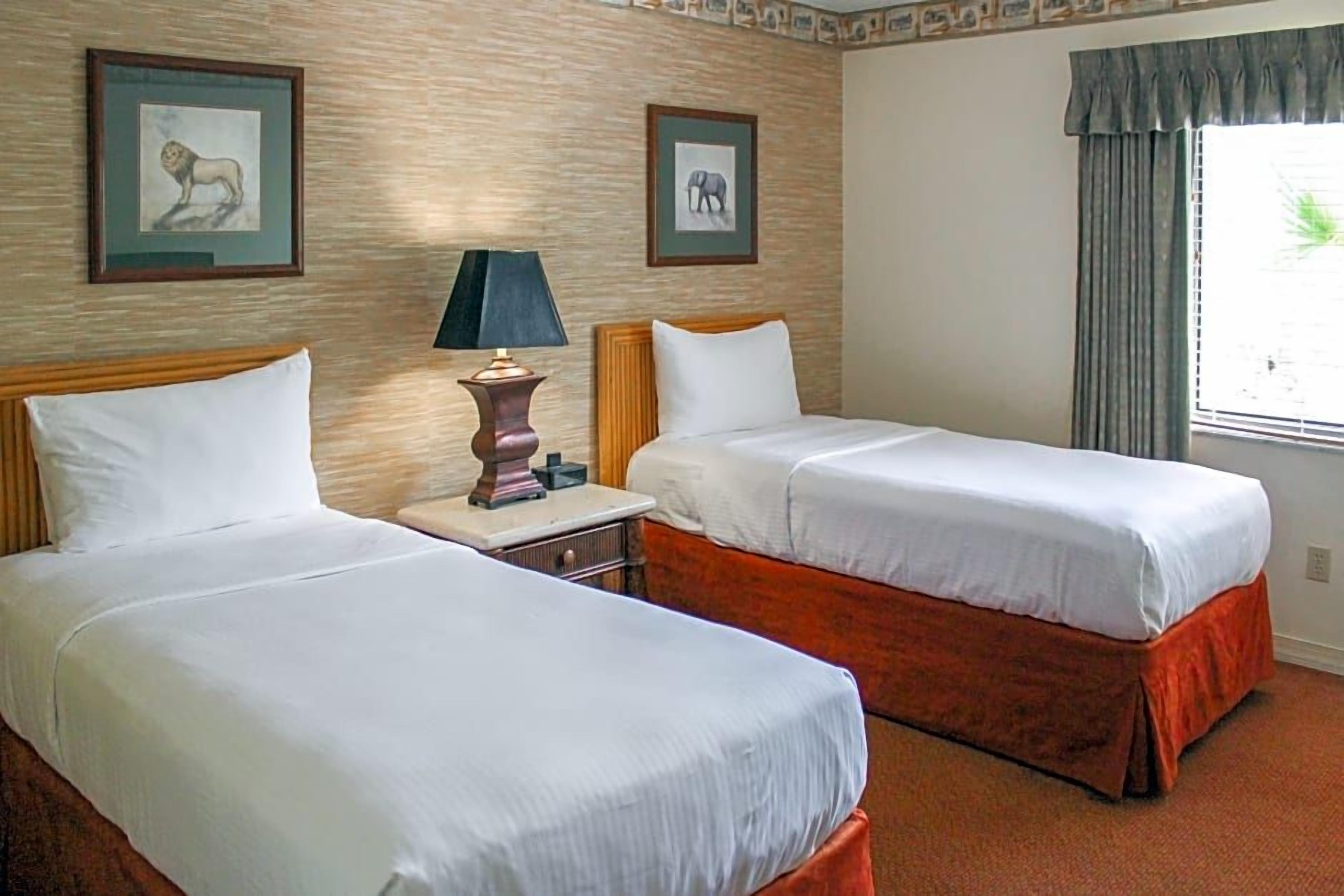 A hotel room with two beds, a nightstand, a lamp, and framed pictures on the wall.