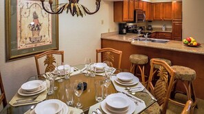 Un coin repas avec une table dressée pour six personnes, des chaises en bois, un lustre et une cuisine en arrière-plan.