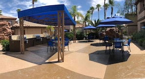 An outdoor patio area with covered seating, a barbecue grill, and palm trees in the background. - Las Vegas | Friends Vegas Retreat | Pool + Hot Tub | 2BR (Las Vegas)