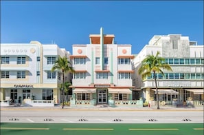 A mid-20th century hotel with a central entrance, palm trees, and adjacent shops.