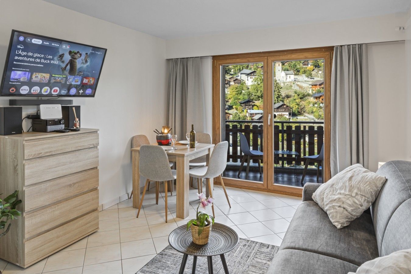 A modern living room with a flat-screen TV, a dining area with a table and chairs, a sofa, and a balcony with a view of houses and greenery.
