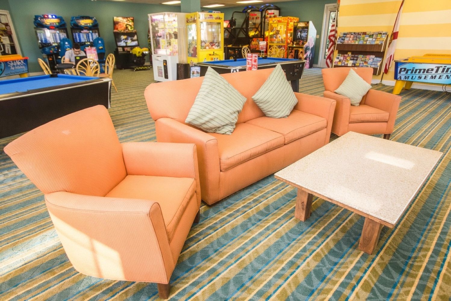 A pool table, orange sofas, and a small table in a room with arcade games in the background.