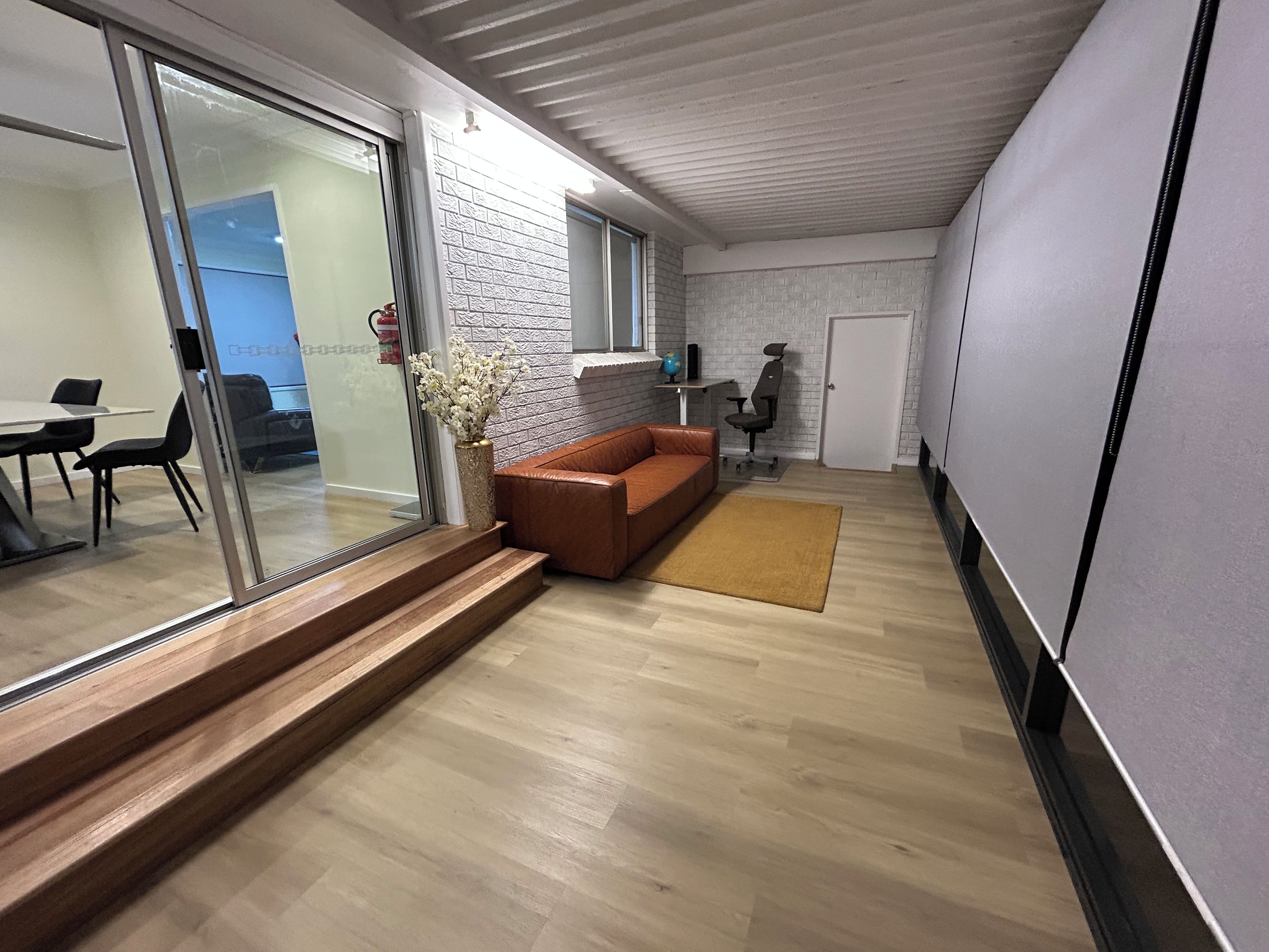 A modern living space with a wooden floor, a brown leather sofa, a glass door leading to a dining area, and a brick wall.