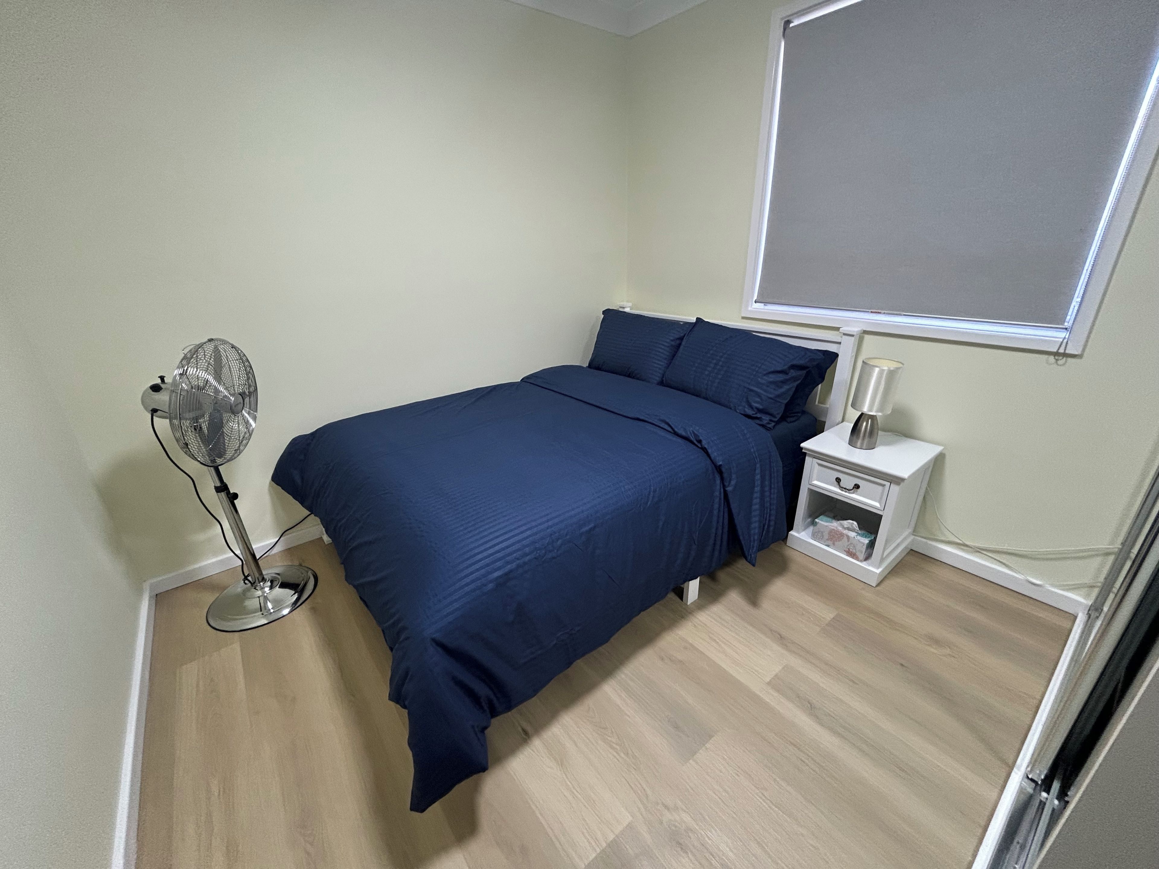 A bedroom with a bed, a bedside table, a nightstand, a fan, and a window.