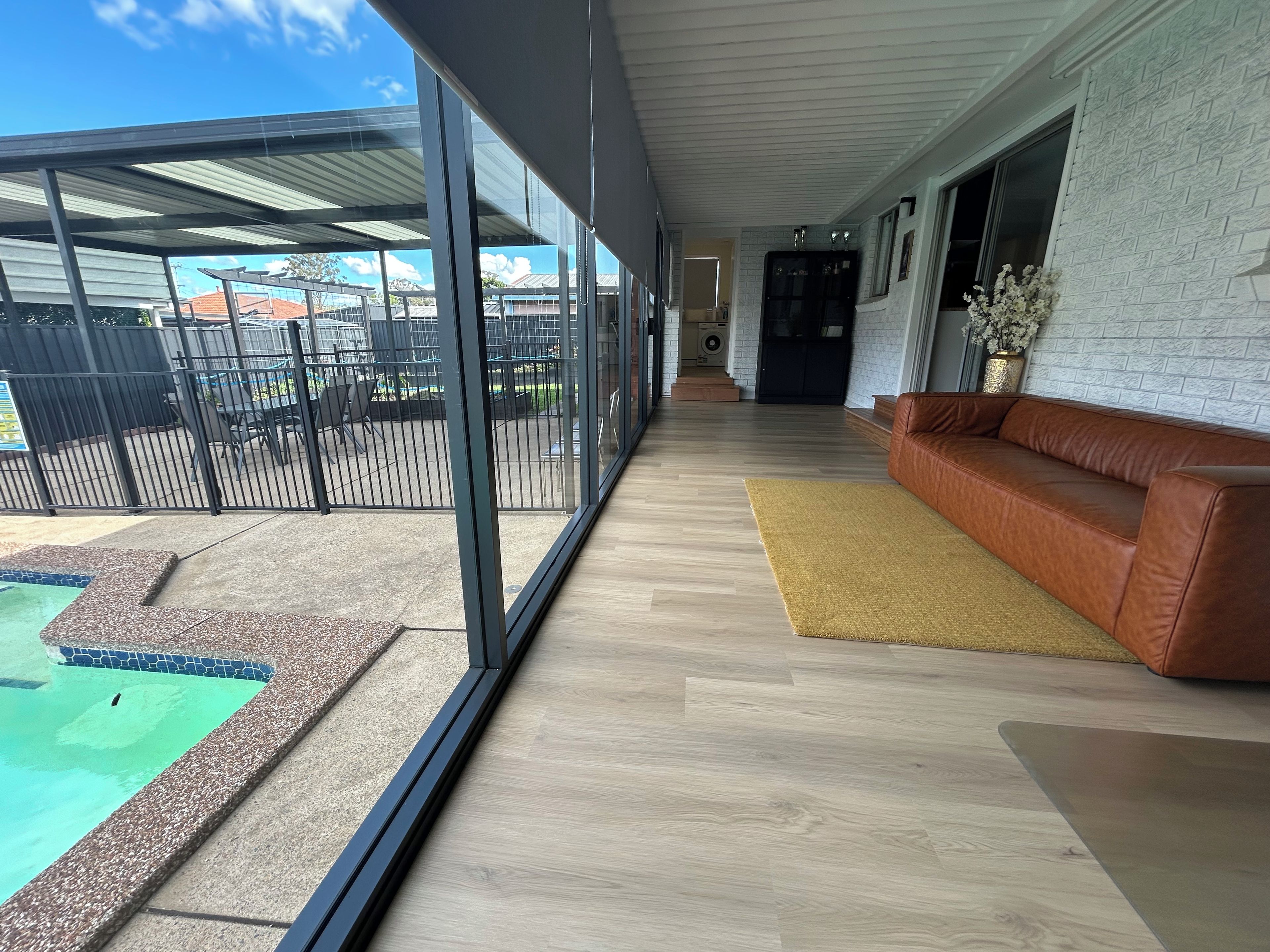 A modern interior with a wooden floor, a brown leather sofa, a glass door leading outside, and a pool area visible through the glass.