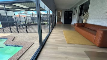 A modern interior with a wooden floor, a brown leather sofa, a glass door leading outside, and a pool area visible through the glass.