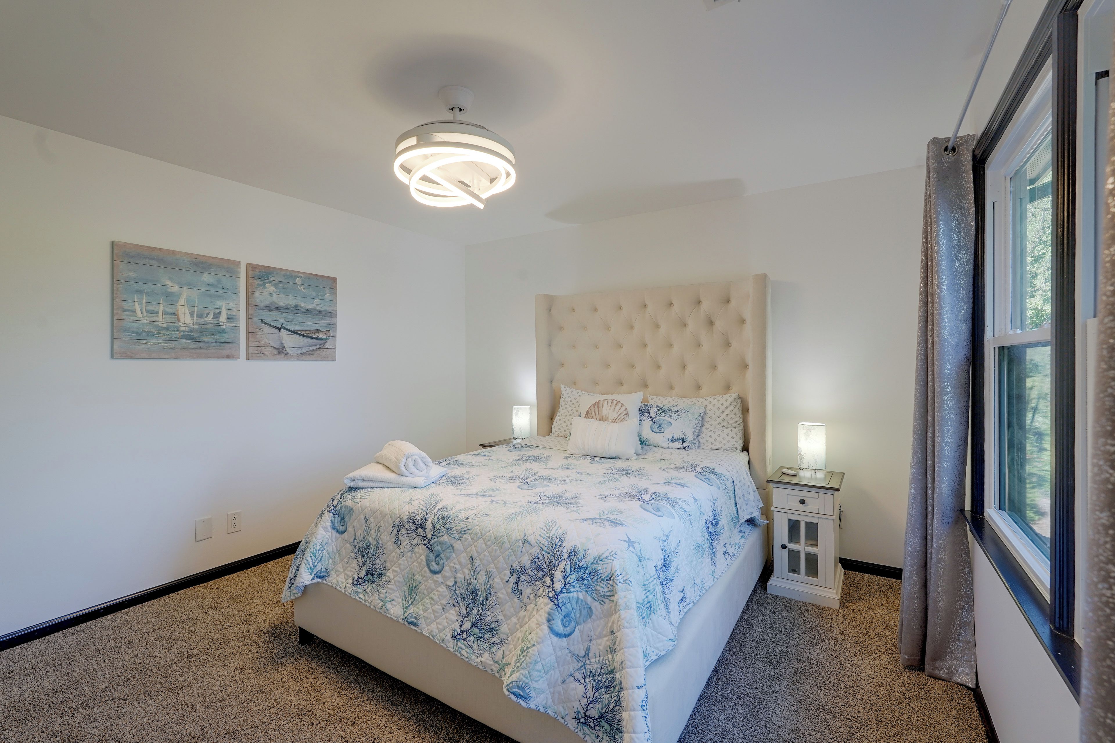 Un dormitorio con cabecera capitonada, cama con colcha estampada de flores azules, dos cuadros en la pared, una lámpara de noche y una ventana con cortinas.