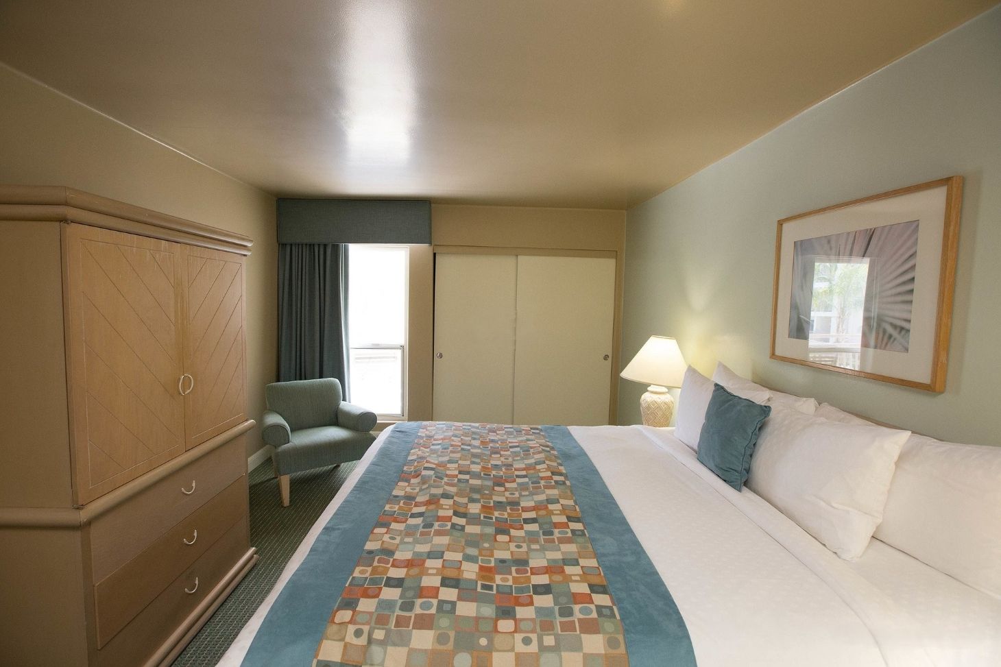 Ein Hotelzimmer mit Bett, Sessel, Kleiderschrank, Tür, Fenster und Lampe.