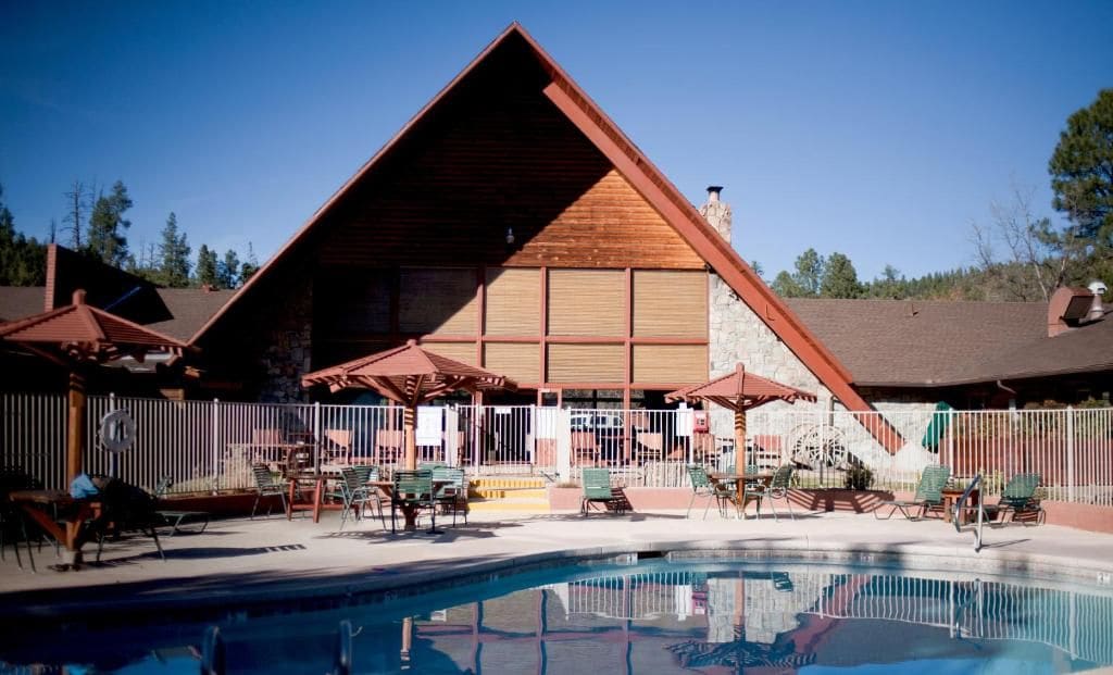 A pool area with lounge chairs and umbrellas, a building with a sloped roof, and a fenced perimeter.