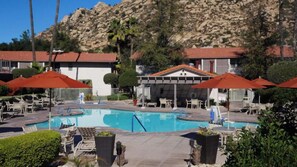 A pool area with lounge chairs and umbrellas, a pool, and buildings with red roofs in the background.