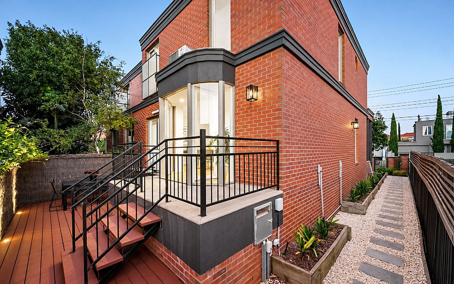 A two-story house with a balcony, a staircase leading to the entrance, and a garden area with plants.