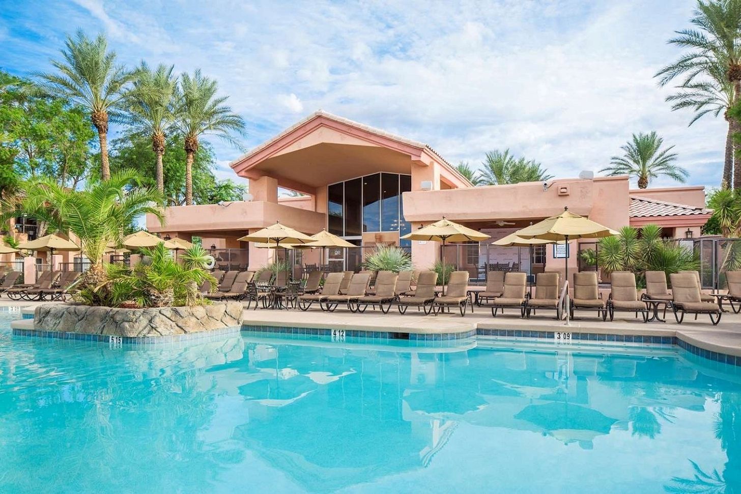 A pool area with lounge chairs and umbrellas, surrounded by palm trees and a building with a large window.