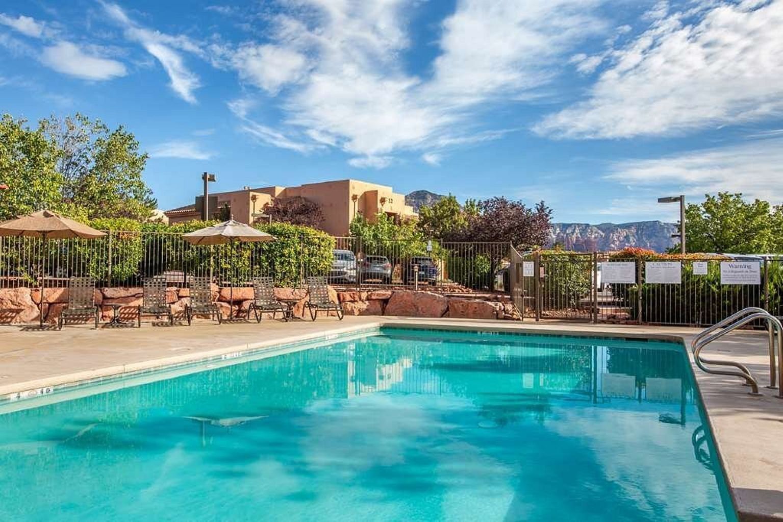 Ein Swimmingpool mit Liegestühlen und Sonnenschirmen, umgeben von einem Zaun und Bäumen.