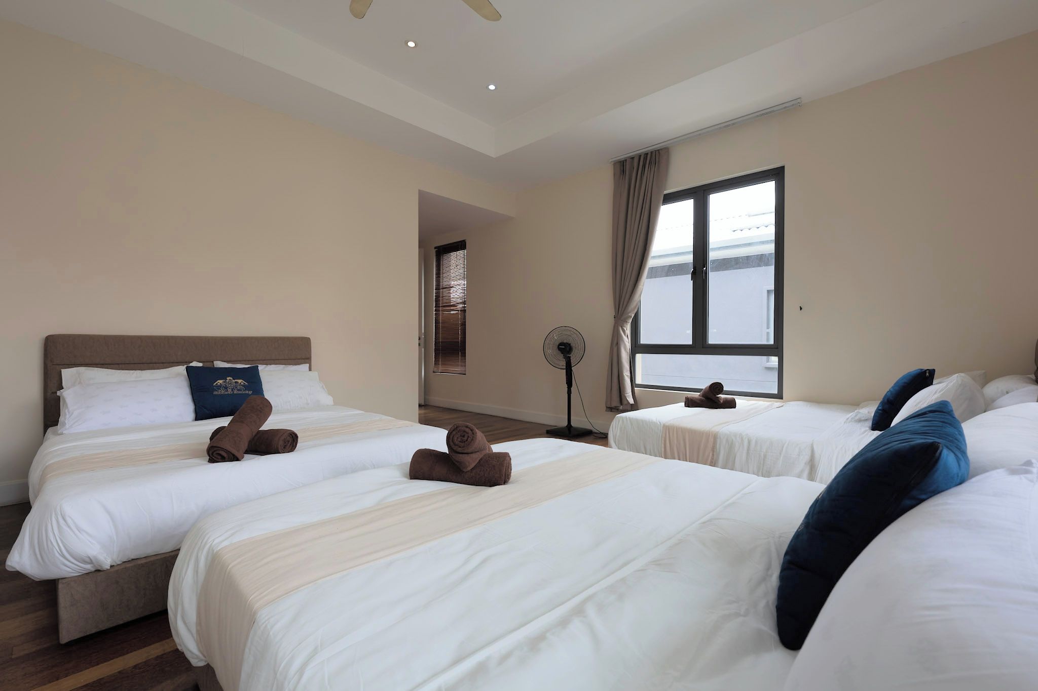 A hotel room with three beds, a standing fan, and a window with curtains.
