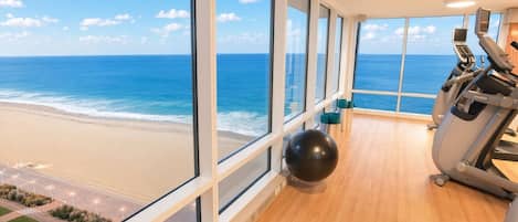 Un gimnasio con equipo de ejercicio y un amplio ventanal con vistas a la playa.