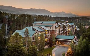 Un hotel de varios pisos con techo verde, rodeado de árboles y montañas al fondo.