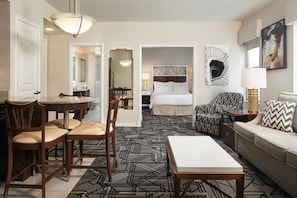 Quarto de hotel com cama, área de estar com sofá e poltronas, mesa de jantar com cadeiras e corredor que dá para um banheiro.