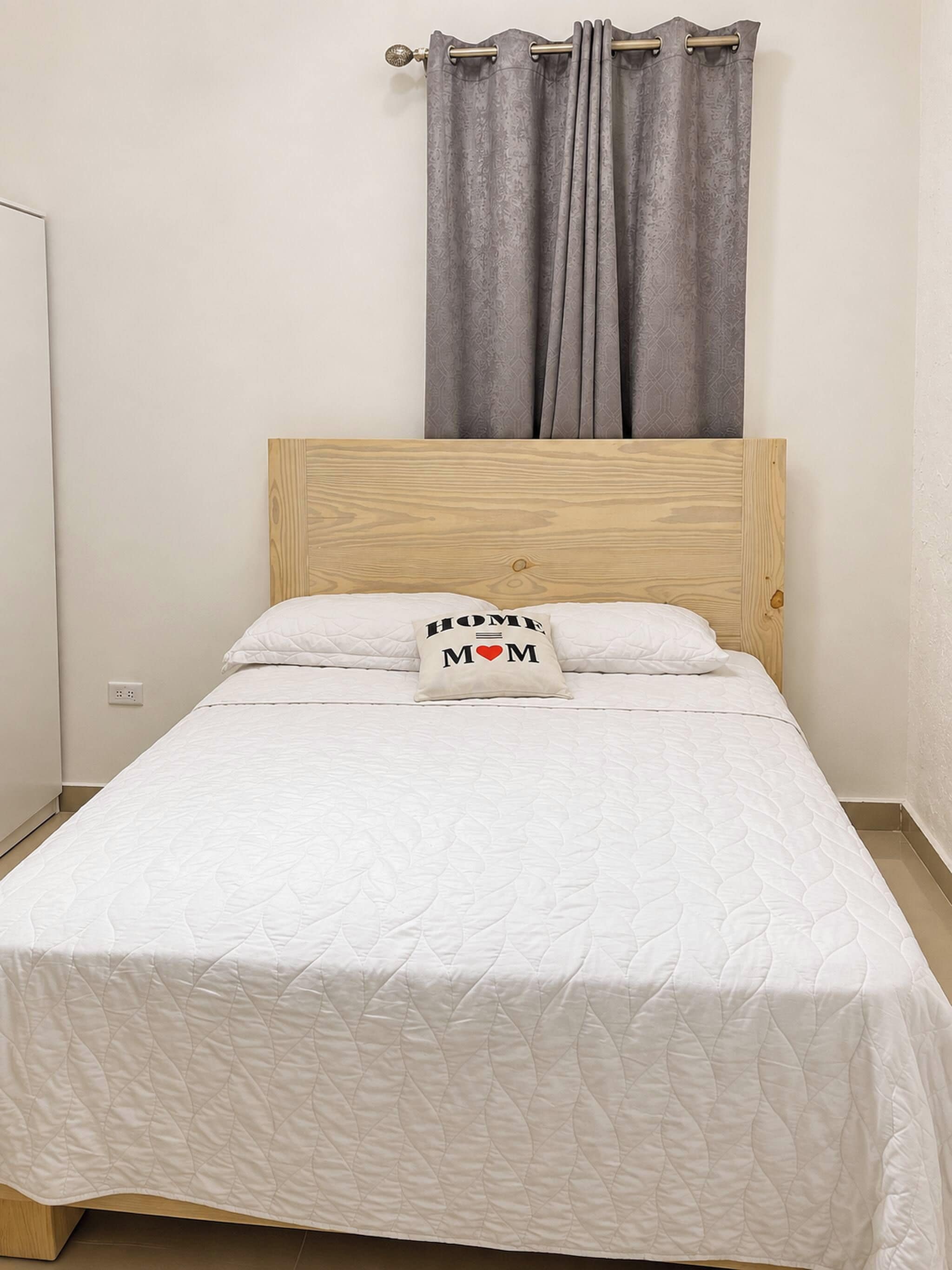 Een eenpersoonsbed met een houten hoofdbord, een witte geborduurde spreideken en een kussen met de tekst 'HOME MOM'.
