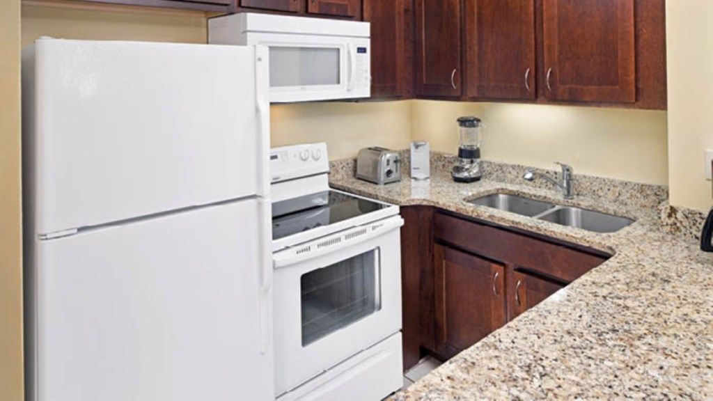 A kitchen with a refrigerator, microwave, toaster, blender, oven, granite countertop, and wooden cabinets.