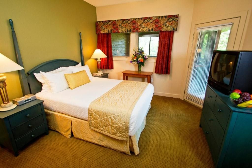 A hotel room with a bed, nightstand, lamp, TV, and a window with curtains.