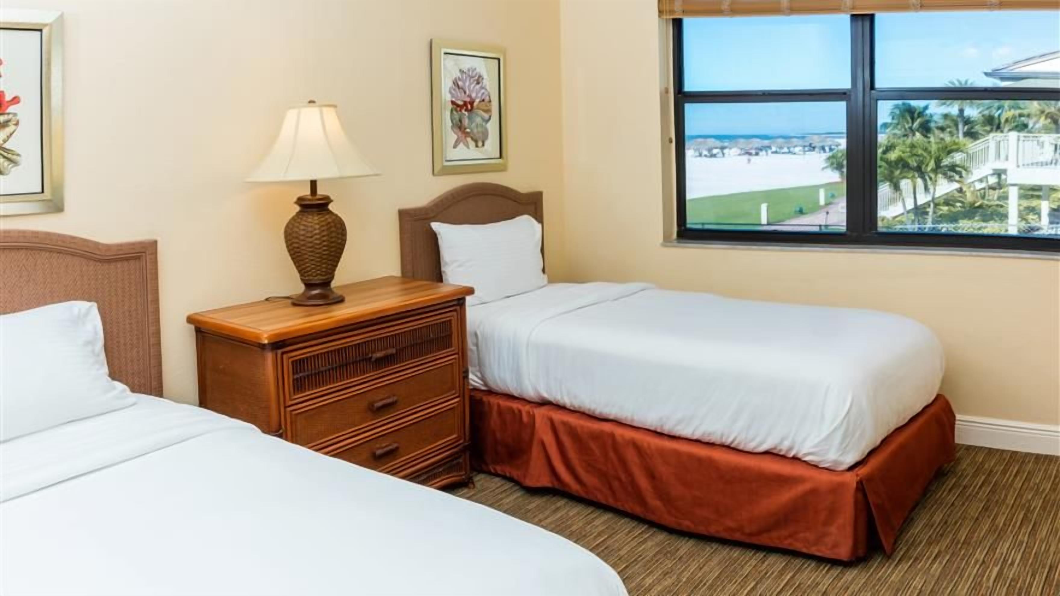 A hotel room with two beds, a nightstand with a lamp, and a window with a view of the beach.