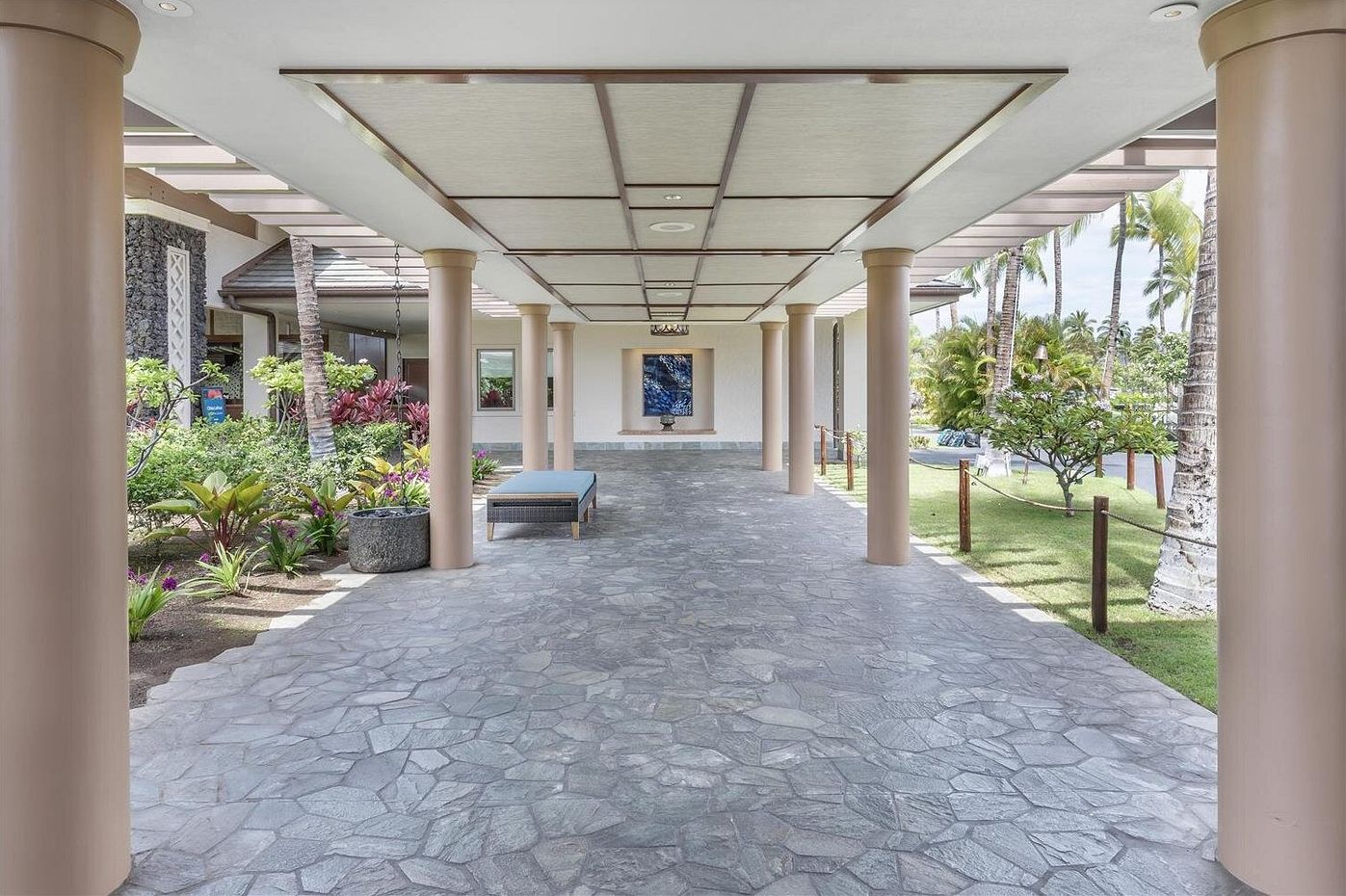 A covered walkway with stone flooring, columns, and a bench.