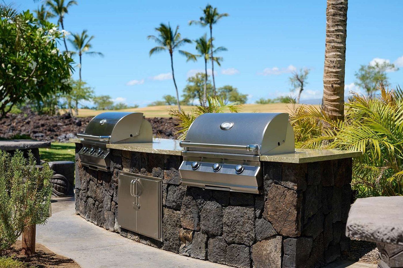 An outdoor grill setup with a stone wall, a storage cabinet, and a clear view of palm trees and a golf course in the background.