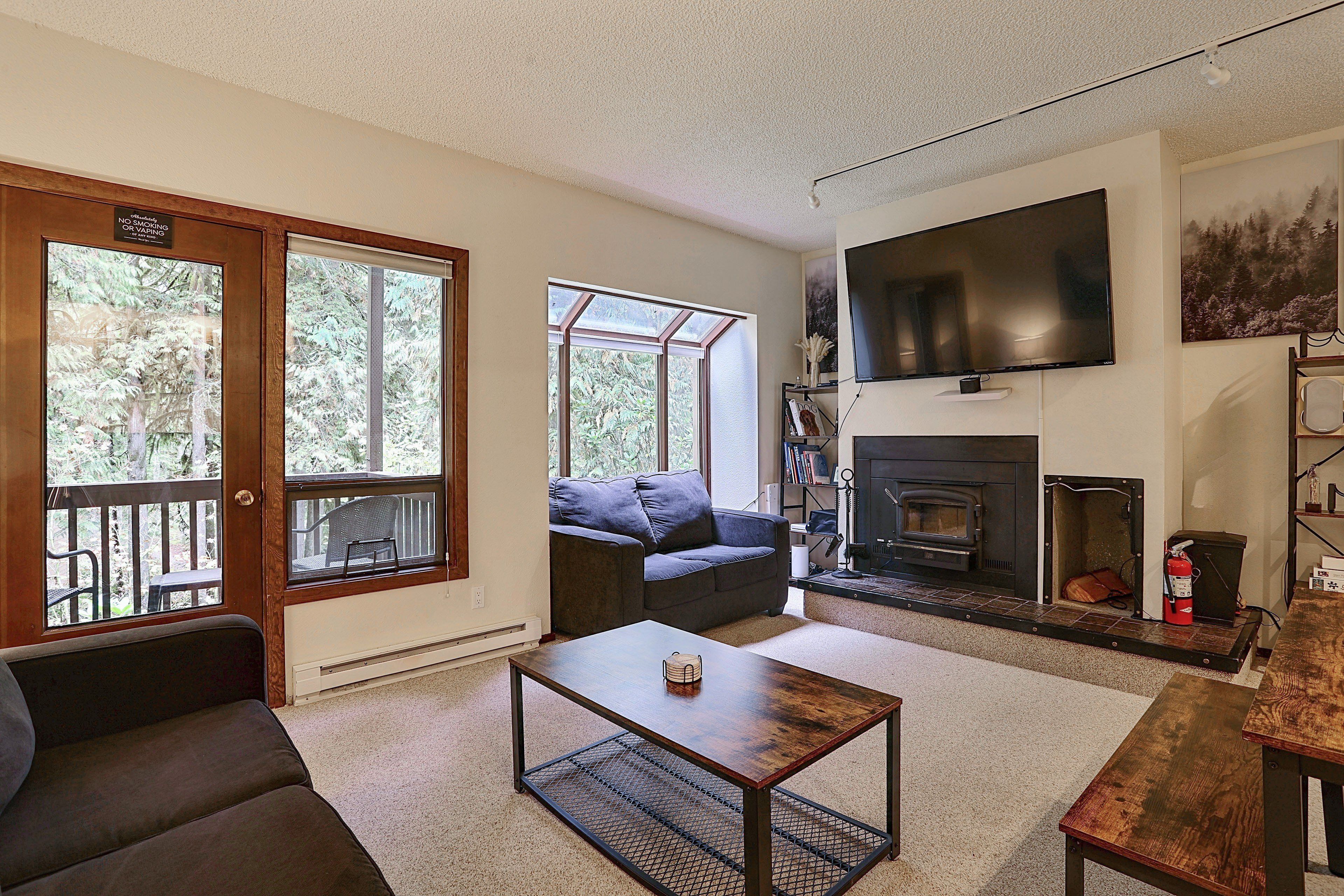A living room with a fireplace, a television mounted above it, and a large window with a view of trees.