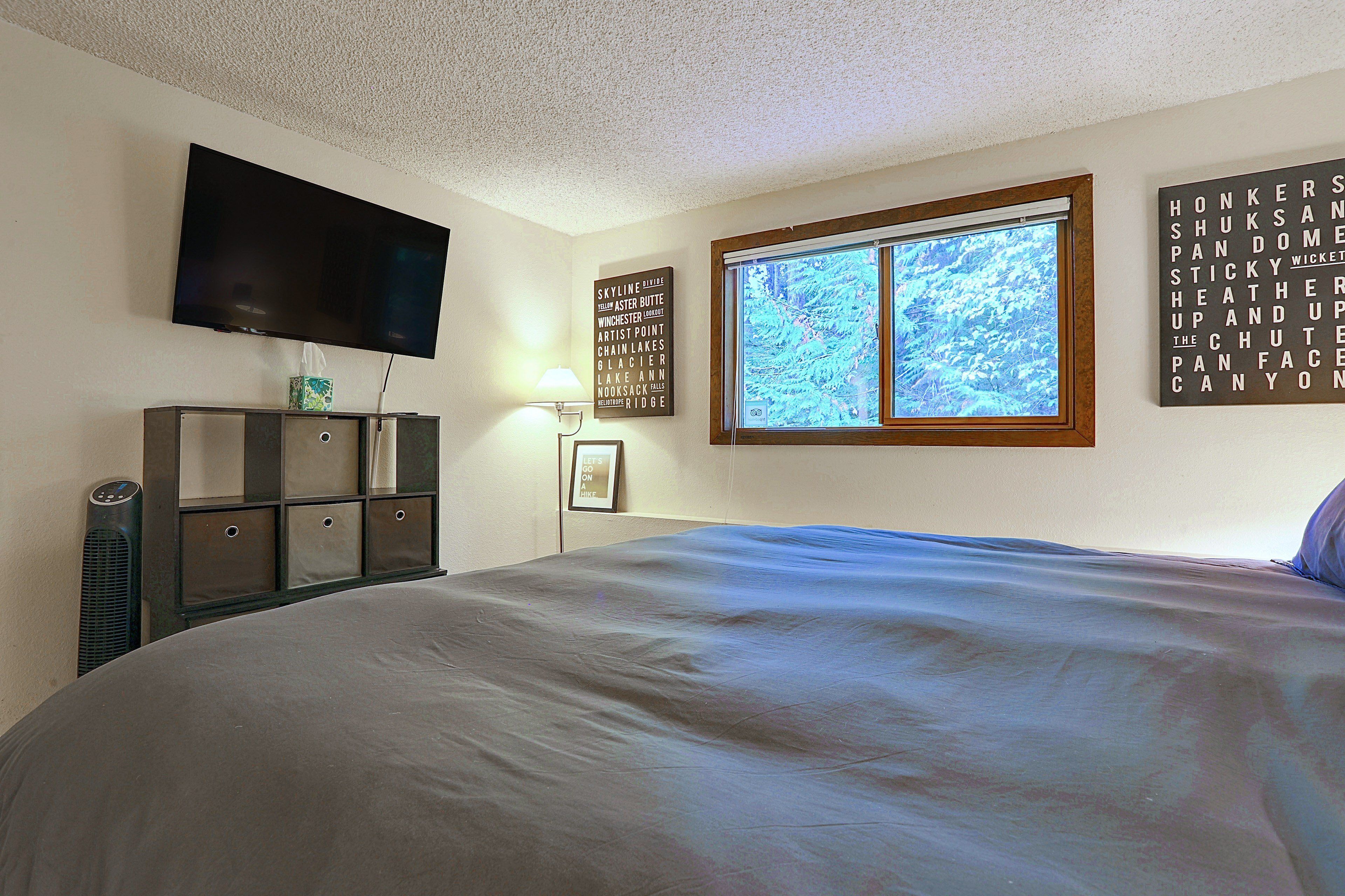 A bedroom with a large bed, a flat-screen TV mounted on the wall, a window with a view of trees, and a wall with a word puzzle.