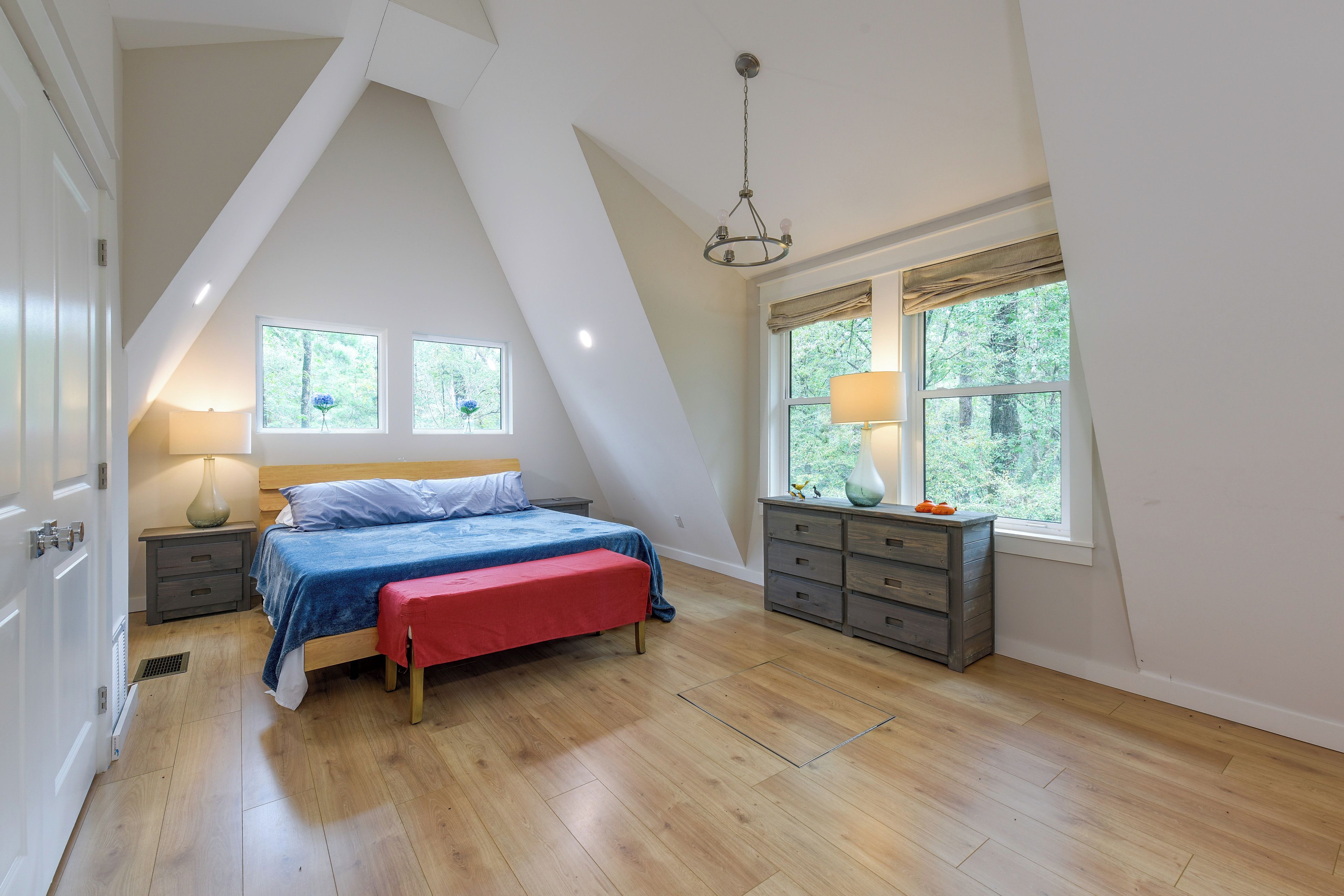 Camera da letto con soffitto spiovente, un letto con una panca rossa, pavimento in legno e due finestre con tende.